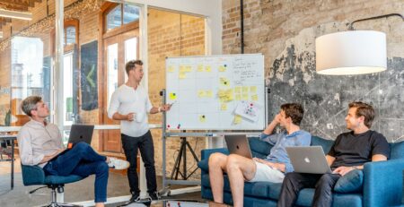 three men sitting while using laptops and watching the man beside the whiteboard