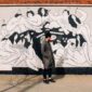 person in a grey coat standing in front of a wall with a mural